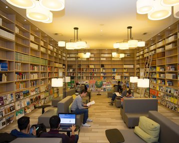 A modern library with shelves full of books. Several people are sitting on sofas, using laptops or reading. Bright ceiling lights create a pleasant atmosphere. A globe stands in the center of the room.