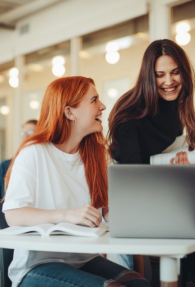 A group of students is sitting in a modern classroom. Two women are laughing and chatting, while a third woman is working on her laptop. Other students in the background are busy with their laptops and notes.