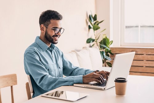 A man with glasses is sitting at a table, working on a laptop. He is wearing a light blue shirt and smiling as he types. On the table, there is a coffee cup and a tablet.