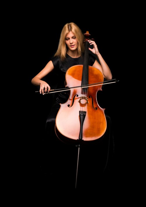 A woman with long blonde hair is playing a cello. She sits in a dimly lit room, holding the bow in her right hand while pressing the strings with her left.