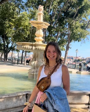 Young woman with brown hair standing in front of a fountain, wearing a white top and a denim jacket, holding a bag in her hand.