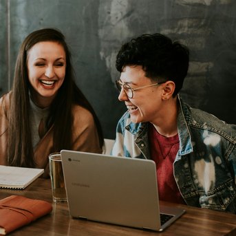 Three people are sitting at a table in a café, working on laptops and laughing. On the table, there’s a notebook and a glass of water. The atmosphere feels relaxed and collegial.
