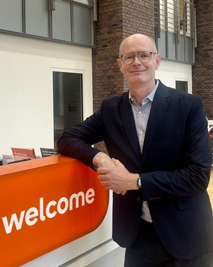 A man with glasses stands at a reception desk with the sign 'welcome.' The background features modern architecture.