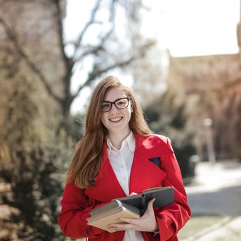 A smiling woman with long red hair is wearing a red jacket and holding several books in her hands. In the background, there are trees and a historic building.