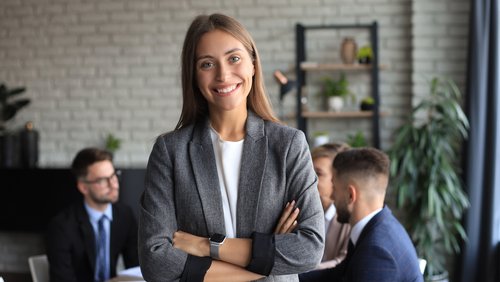 Eine lächelnde Frau in einem grauen Blazer steht im Vordergrund, während im Hintergrund mehrere Personen an einem Tisch diskutieren.