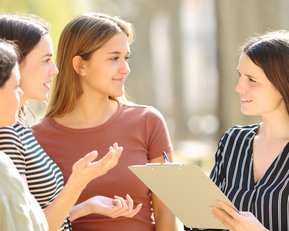 Vier Frauen stehen im Freien und diskutieren. Eine Frau hält ein Clipboard und spricht, während die anderen aufmerksam zuhören.