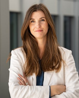 A woman with long brown hair and a white blazer stands with her arms crossed in front of a modern building.
