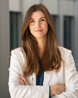 A woman with long brown hair and a white blazer stands with her arms crossed in front of a modern building.