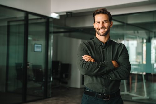 Ein junger Mann mit einem breiten Lächeln steht in einem modernen Büro, die Arme vor der Brust verschränkt, in einem dunkelgrünen Hemd.