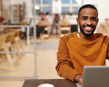 Ein junger Mann mit einem breiten Lächeln sitzt an einem Tisch und arbeitet an einem Laptop. Er trägt einen orangefarbenen Pullover und befindet sich in einem modernen, hellen Raum mit Holzinterieur.