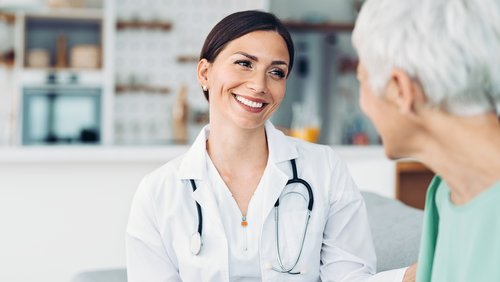 Eine Ärztin in weißem Kittel lächelt und spricht mit einer älteren Patientin in einem hellen, modernen Raum.