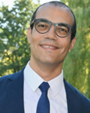 A man in glasses and a suit smiles at the camera, surrounded by green trees.