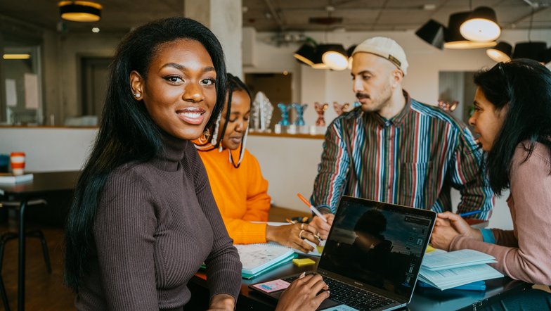 A woman with long, black hair sits at a table, smiling as she looks at a laptop. Around her, three other people are working on notes and projects. The room is modern and bright.