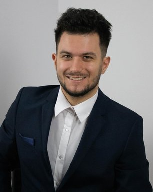 Ein junger Mann in einem Anzug lächelt in die Kamera, mit einem weißen Hemd und einem dunklen Blazer.