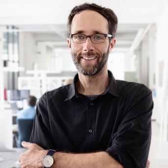 A man with glasses and a beard stands with his arms crossed in a modern office. He is wearing a black shirt and a wristwatch.