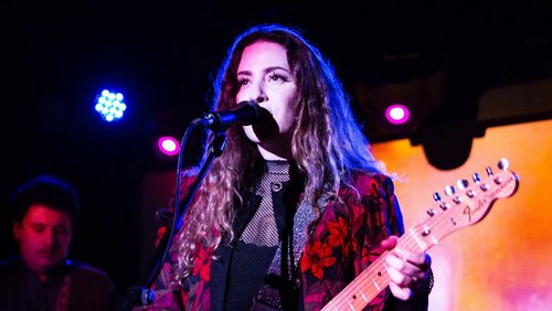 A musician with curly hair stands at the microphone playing guitar. She is wearing a black top with a floral pattern. In the background, lights and another musician can be seen.