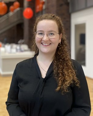 A smiling woman with curly brown hair and glasses is wearing a black top. In the background, blurred red decorations are visible.