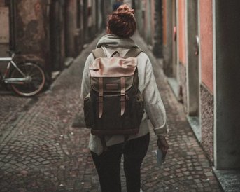 Junge Studentin mit Rucksack von hinten.  A person with a backpack walks down a narrow, cobblestone alley. In the background, there are old buildings and a bicycle. The atmosphere feels calm and nostalgic.