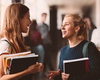 Two students are chatting in a hallway while holding books and notes in their hands. In the background, other students can be seen moving around the school.