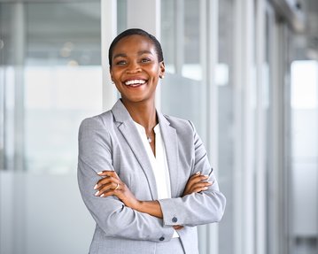 A smiling woman in a gray suit stands with her arms crossed in a modern office.