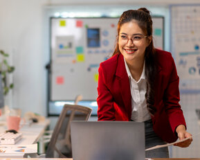 Eine Frau in einem roten Blazer steht an einem Schreibtisch, lächelt und arbeitet an einem Laptop, umgeben von Dokumenten und Büromaterialien.