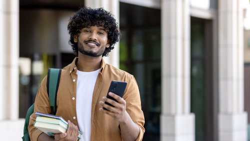 A young man with curly hair is holding a smartphone in one hand and books under his arm while smiling. He is wearing a backpack.