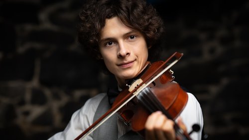 A young man with curly hair is playing the violin. He is wearing a white shirt and a gray vest while looking at the camera. The background is blurred and features dark stones.