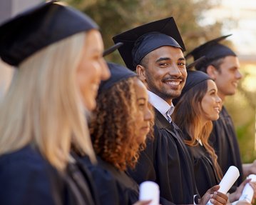 Sechs Absolventen in schwarzer Abschlusskleidung stehen in einer Reihe, halten ihre Diplome in den Händen und lächeln. Die Gruppe zeigt eine Vielfalt an Haarstilen und Geschlechtern. Der Hintergrund ist unscharf und zeigt eine akademische Umgebung.