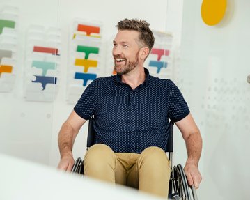 Ein Mann mit Bart sitzt in einem Rollstuhl und lächelt. Im Hintergrund sind bunte Farbmuster sichtbar.