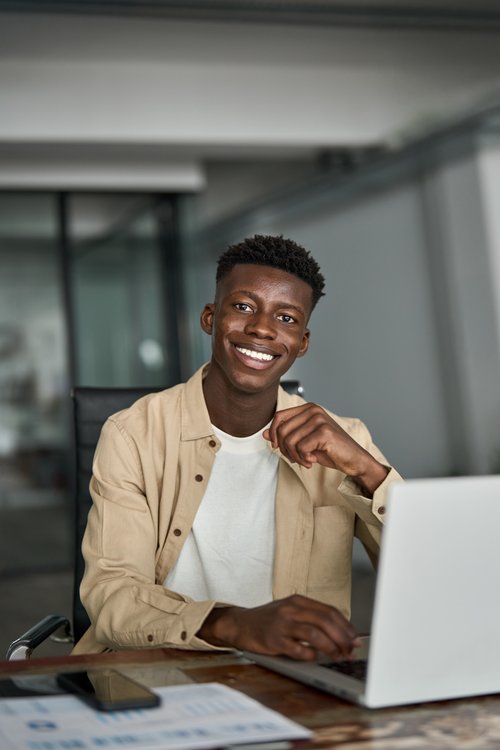A young man is sitting at a desk, smiling and working on a laptop. On the table, there is a smartphone and some documents.