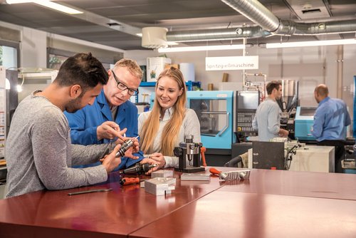 Two people are discussing a mechanical part with a teacher at a table in a technical workshop. Machines are visible in the background.