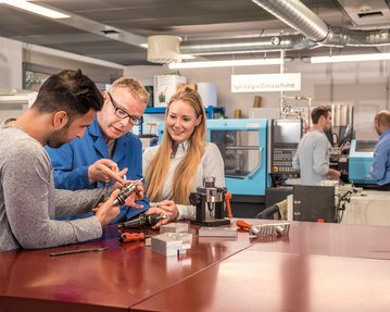 Two people are discussing a mechanical part with a teacher at a table in a technical workshop. Machines are visible in the background.