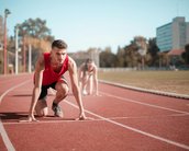 A young athlete in a red jersey is in the starting position on a track. In the background, another runner is also getting ready to start. The setting features a sports facility with trees and buildings.