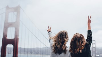 Zwei Personen stehen mit dem Rücken zur Kamera und zeigen mit beiden Händen das Peace-Zeichen. Im Hintergrund ist die Golden Gate Bridge und ein bewölkter Himmel zu sehen.