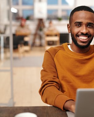 A young man with a broad smile sits at a table, working on a laptop. He is wearing an orange sweater and is in a modern, bright room with wooden interiors.