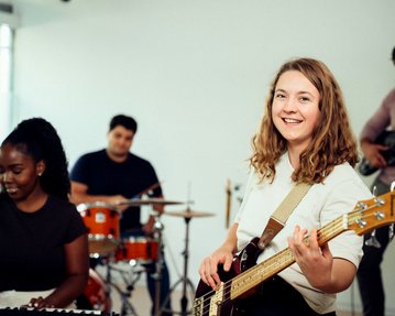 Eine Frau mit einer Bassgitarre lächelt in die Kamera, während sie in einer Bandprobe mit anderen Musikern spielt.
