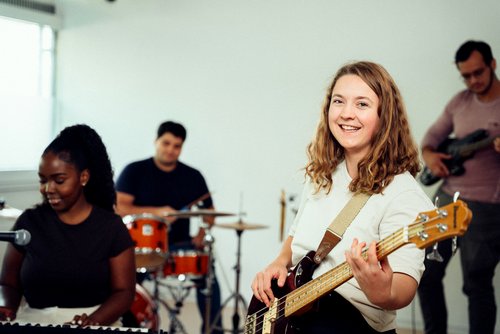 A woman with a bass guitar smiles at the camera while playing in a band rehearsal with other musicians.