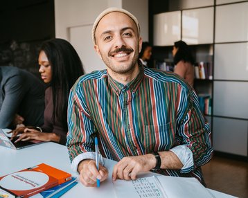 A man in a striped shirt and a cap sits at a table, smiling as he writes with a pen on a notepad. In the background, there are other people with laptops and books visible.