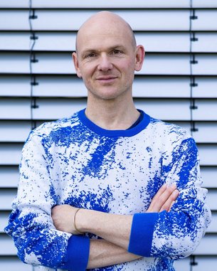 A bald man wearing a blue and white sweater stands with his arms crossed in front of a striped wall.