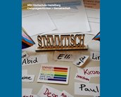 Im Mittelpunkt steht ein Holzschild auf dem Tisch: „Stammtisch“. Hinter dem Schild liegen drei beschriebene Zettel, vor dem Schild sammeln sich verschiedene Namen mit unterschiedlichen Pronomen und ein Sticker in Regenbogenfarben. Einer der Namen ist „Elli“ mit sie/ihr Pronomen. Der Text „SRH Hochschule Heidelberg Campusgeschichten > Gemeinschaft“ steht am oberen Rand.