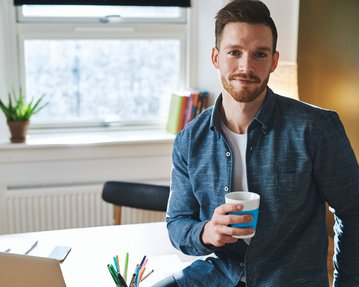Ein Mann mit einem Bart hält einen Kaffeebecher in der Hand. Er trägt ein blaues Hemd und sitzt an einem Schreibtisch mit einem Laptop und Schreibutensilien. Im Hintergrund sind Pflanzen und Bücherregale sichtbar.