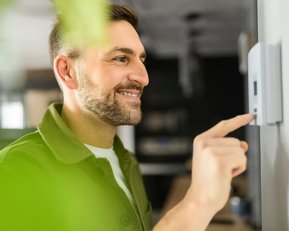 Ein Mann stellt einen Thermostat ein und lächelt dabei. Im Hintergrund sind unscharfe Möbel und Pflanzen sichtbar.