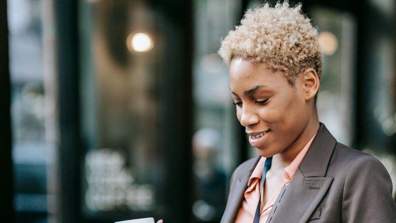 A woman with short, curly hair is sitting at a table, working on a laptop. She is holding a coffee mug in one hand and is wearing a blazer along with an ID badge around her neck.