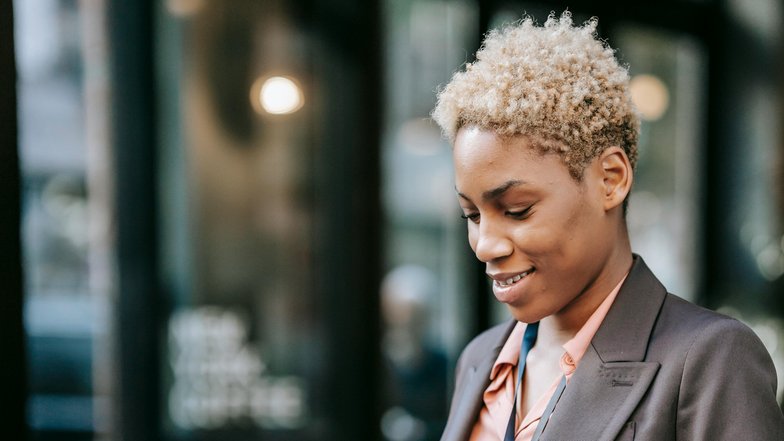 Eine Frau mit kurzen, lockigen Haaren sitzt an einem Tisch und arbeitet an einem Laptop. Sie hält einen Kaffeebecher in der Hand und trägt einen Blazer sowie eine ID-Karte um den Hals.