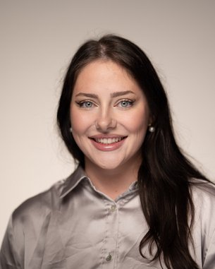 A smiling woman with long, dark hair is wearing a gray shirt and standing against a neutral background.