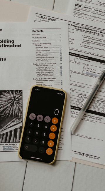 A hand is holding a check, while a smartphone with a calculator is visible. Surrounded by tax documents, including forms for tax deductions and estimated taxes for the year 2019, as well as a pen.