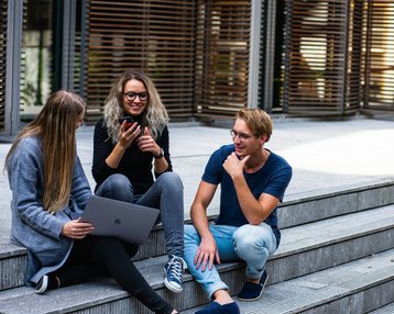 Three people are sitting on steps and interacting with each other. One person is using a laptop while the others are looking at a smartphone. They are all dressed casually and appear to be in an urban setting.