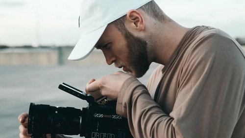 A man wearing a white cap is carefully examining a camera. He holds the camera with one hand and adjusts it with the other. The emphasis is on his concentration and the professional equipment.