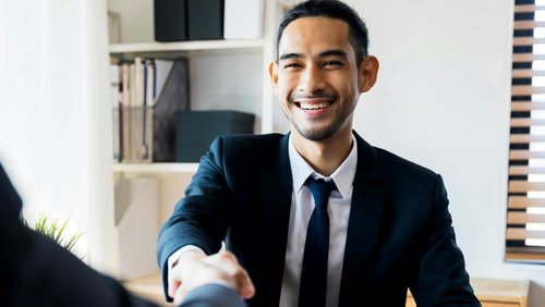 Ein Mann in einem Anzug schüttelt einer anderen Person die Hand und lächelt dabei, während er an einem Tisch sitzt.