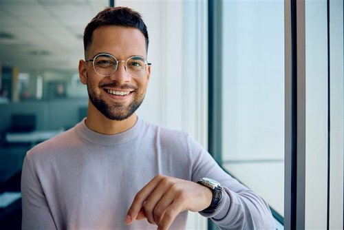 A young man with glasses and a beard smiles as he stands by a window. He is wearing a light sweater and has a watch on his wrist.